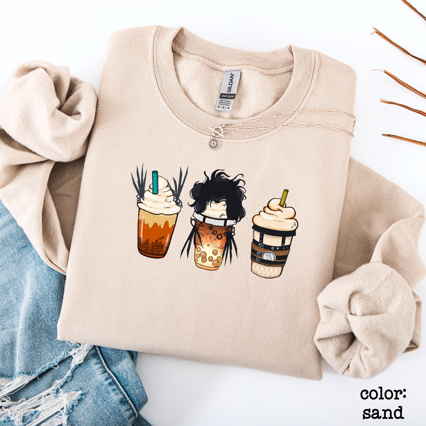Flat lay of a sand-colored graphic sweatshirt featuring three spooky Halloween-themed iced coffee cups side-by-side with funny character details.